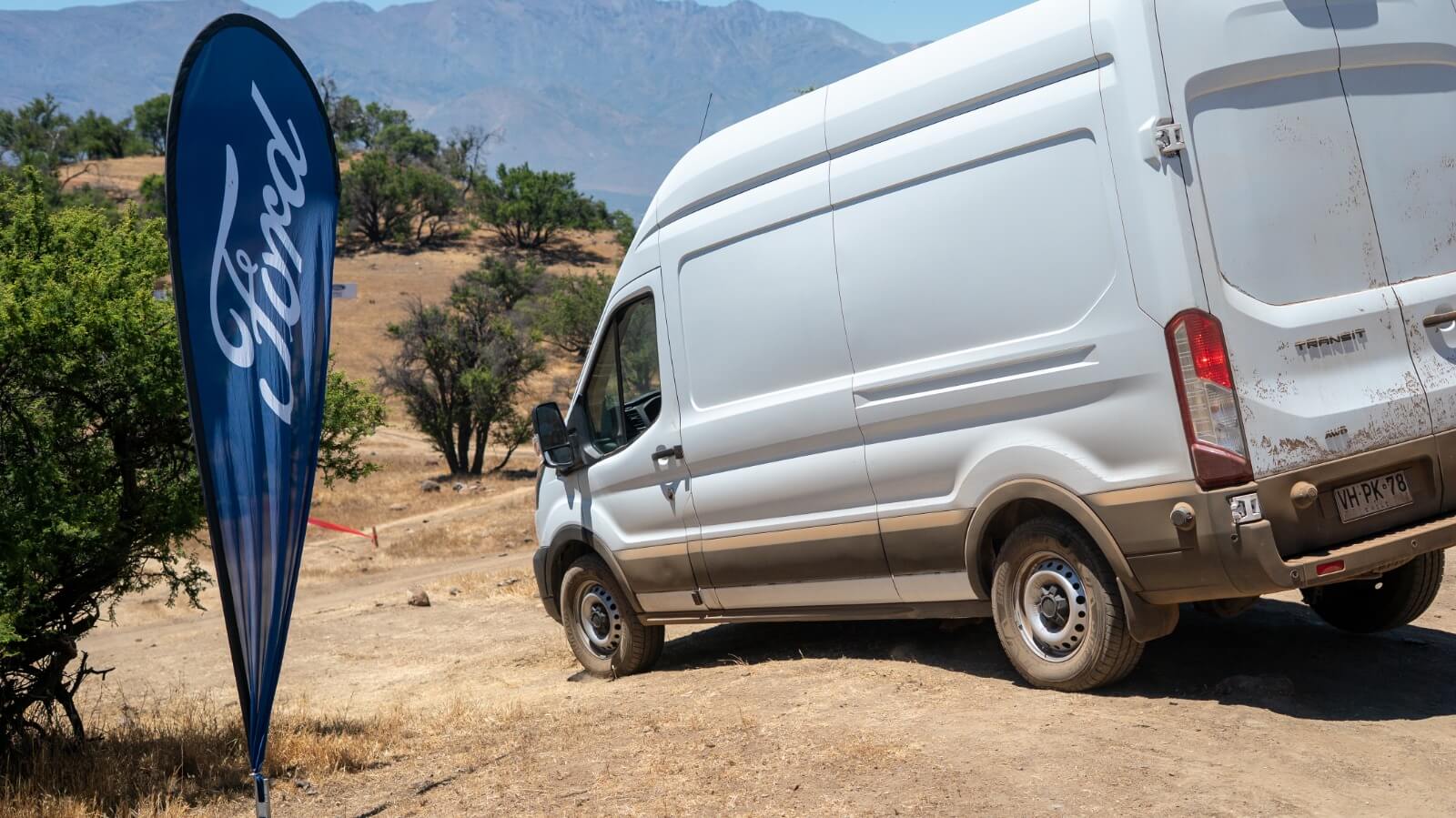 Parte trasera de una Ford Transit blanca sucia en un camino de tierra, con un banderín azul de Ford y montañas de fondo.